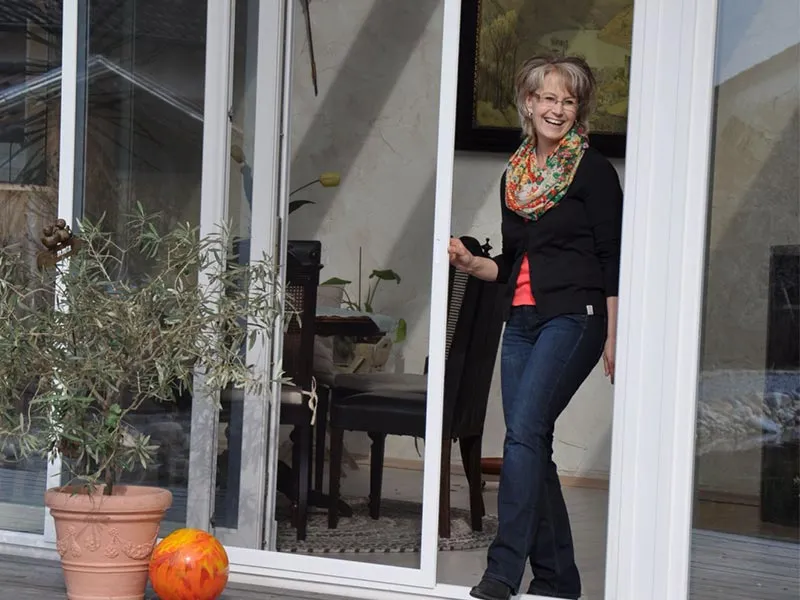 Huber Franz - Insektenschutz - Fenster - Türen – eine Frau öffnet lachend die Schiebetür zu ihrem Garten